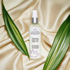 Bottle of Glimmer Goddess organic silky dry body oil on a beige fabric background with green leaves.