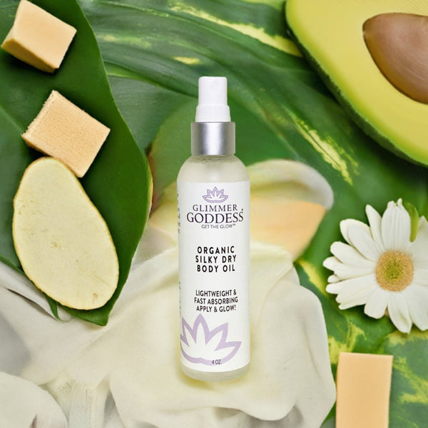 Glimmer Goddess organic silky dry body oil bottle on a green leafy background with avocado and flowers.