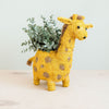 Yellow giraffe-shaped planter with a plant on a light background