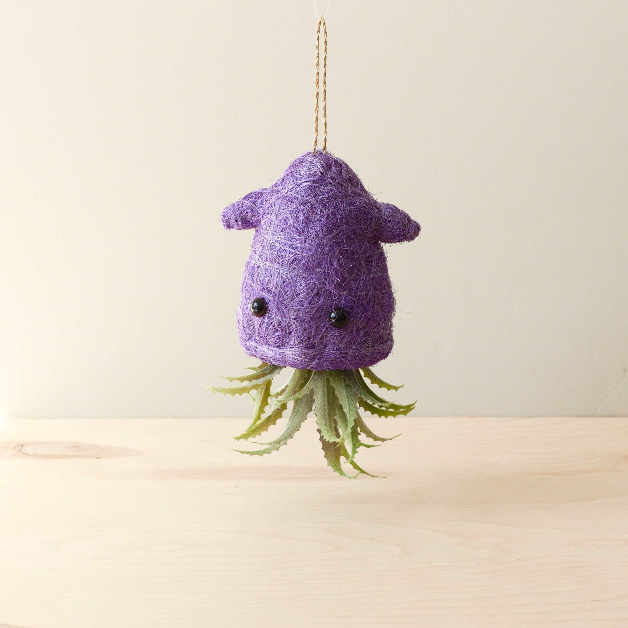 Purple felted creature with black eyes and green plant-like appendages hanging on a beige background