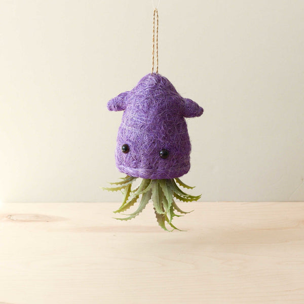 Purple felted creature with black eyes and green plant-like appendages hanging on a beige background