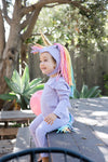 Child wearing a unicorn costume with a rainbow mane sitting on a wooden bench outdoors.