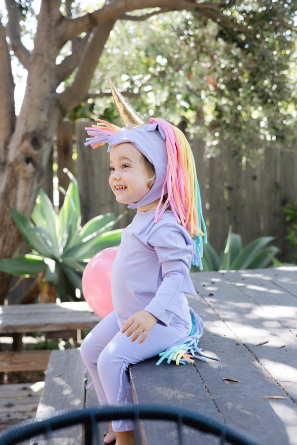 Child wearing a unicorn costume with a rainbow mane sitting on a wooden bench outdoors.