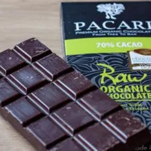 Block of dark chocolate next to a Pacari Raw Organic Chocolate box on a wooden surface. Discover the exquisite taste of Pacari's Raw Unroasted Organic Chocolate Bar. This 70% cacao bar, made with Fair Trade and Vegan Arriba Nacional beans, is the perfect Christmas gift for any chocolate connoisseur.