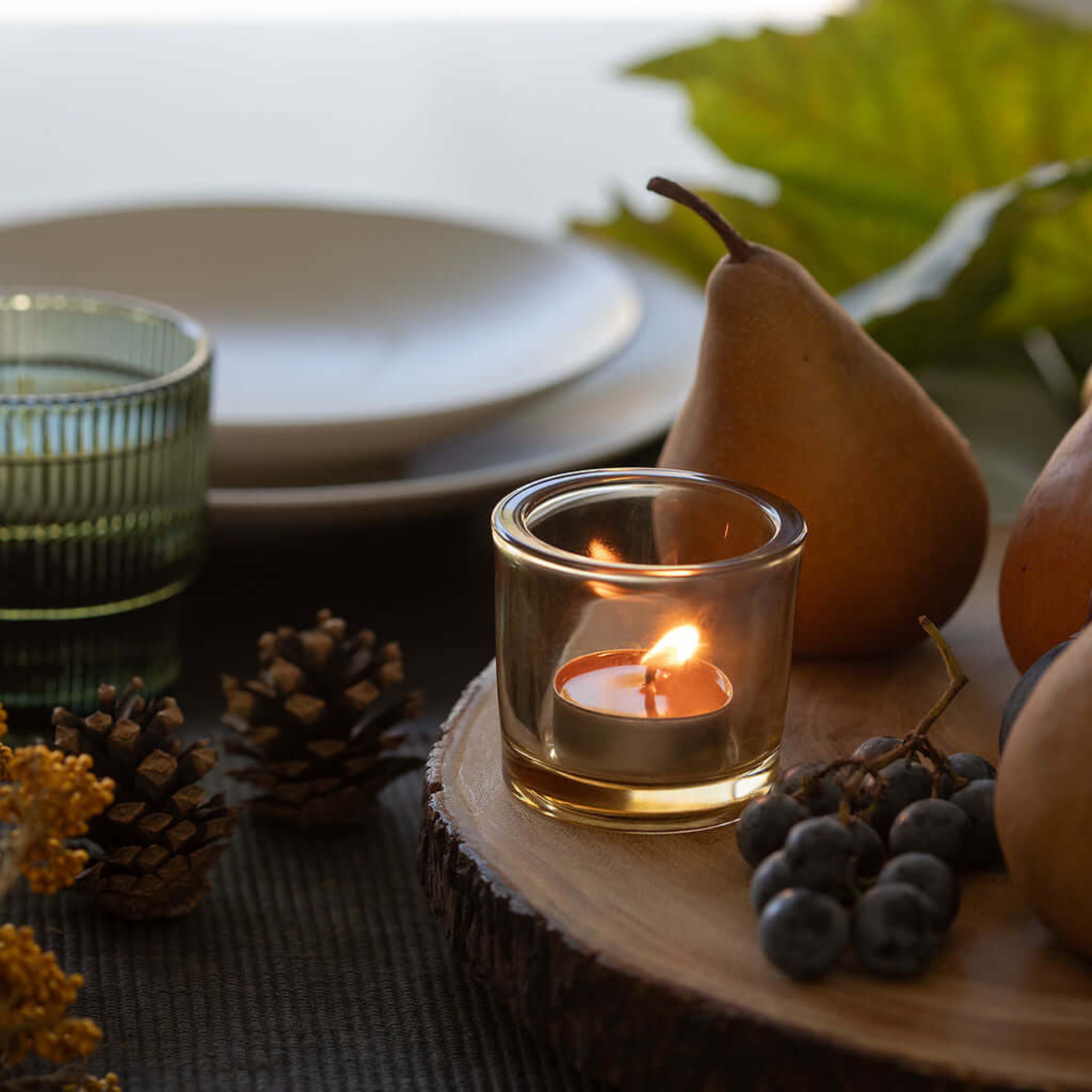 Candle in a small glass holder on a wooden board with fruits and leaves.