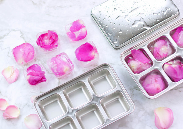 Stainless steel ice cube tray filled with large clear ice cubes next to a whiskey glass