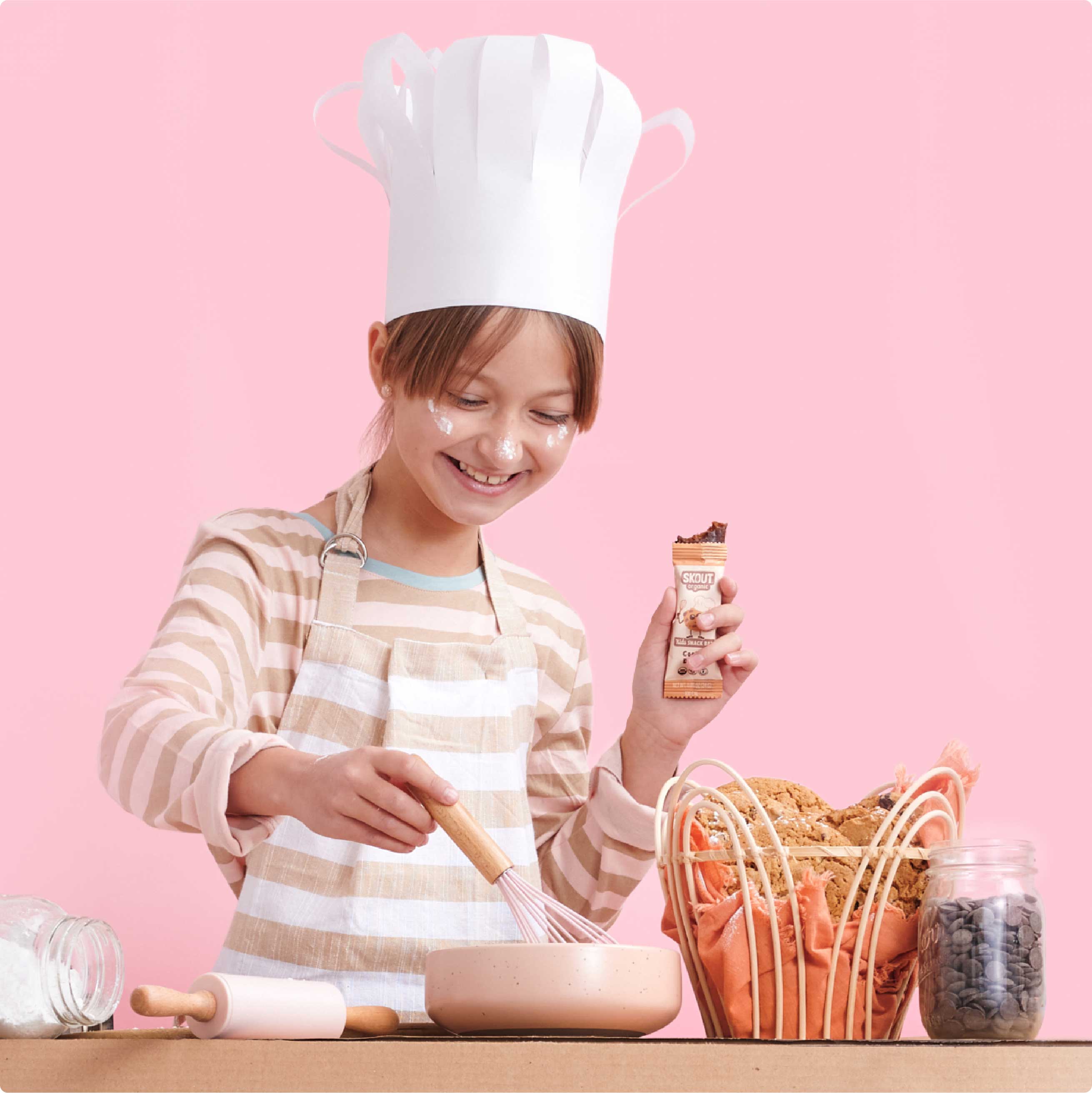 Child in chef's hat and apron holding a chocolate bar with baking items on a pink background