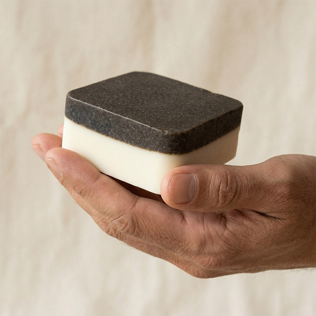 Hand holding a square object with a textured top and smooth bottom against a neutral background