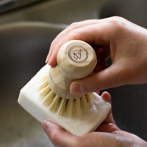 Hand holding a wooden dish brush with a brand logo against a blurred kitchen background. Go plastic-free with the Eco-Friendly Dish Washing Starter Pack. Includes bamboo brush, soap dish, and natural dish soap bar for a sustainable clean.