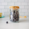 Glass jar with wooden lid containing gray tablets on a white surface with a tiled wall background