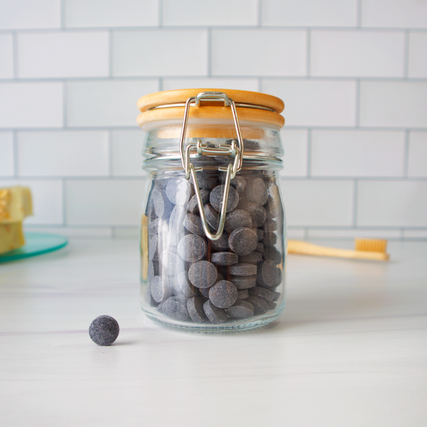 Glass jar with wooden lid containing gray tablets on a white surface with a tiled wall background