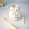 Clear glass jar with white tablets and a wooden toothbrush on a light surface.