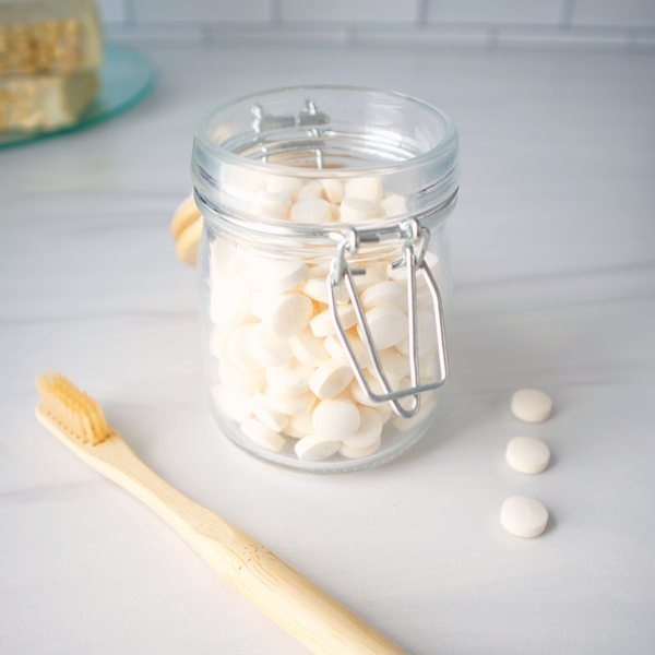 Clear glass jar with white tablets and a wooden toothbrush on a light surface.