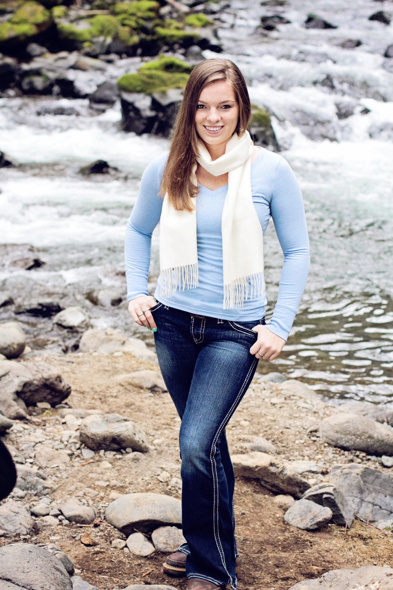 Woman standing by a river wearing a light blue sweater, dark jeans, and a white scarf.