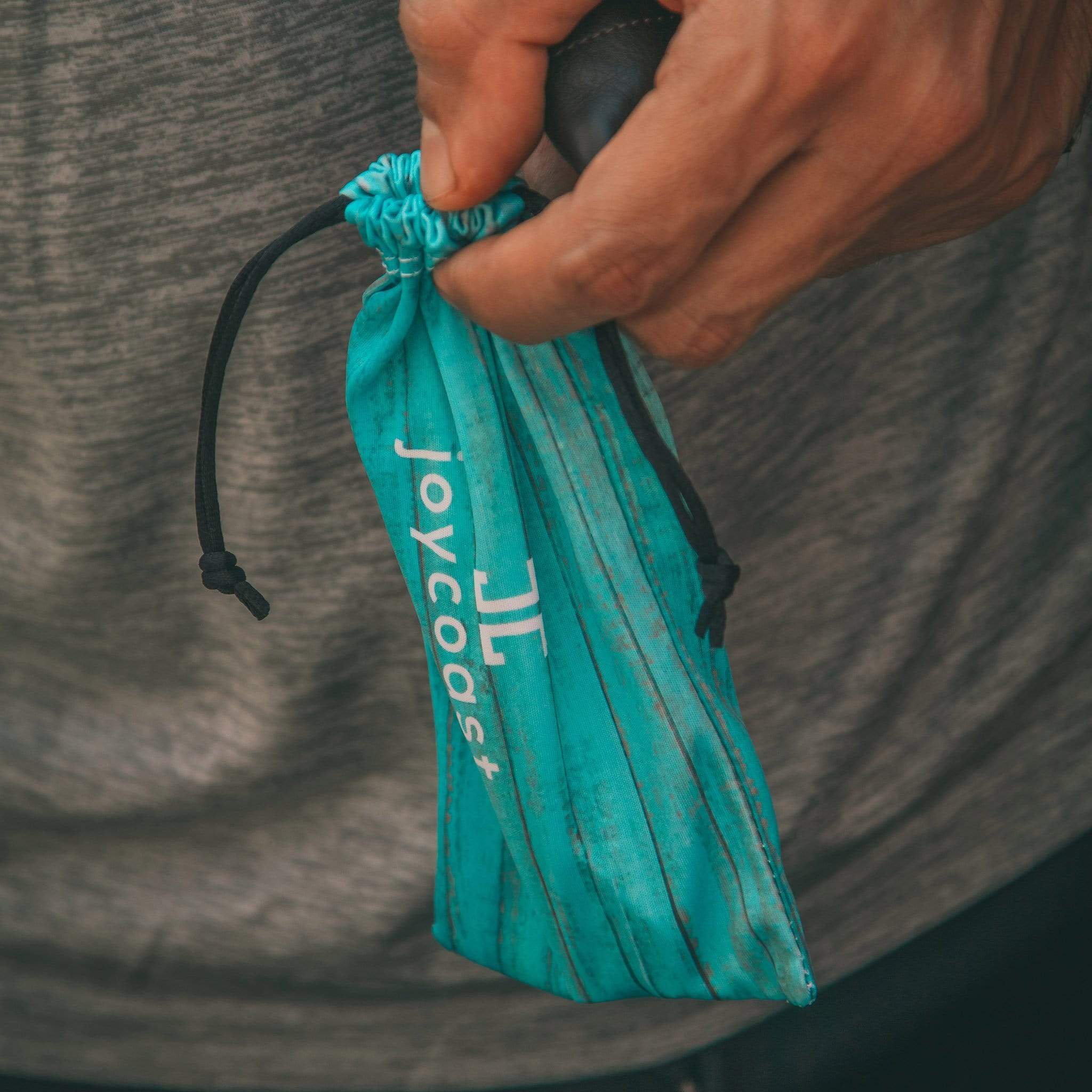 Hand holding a teal drawstring bag with 'joycoast' branding against a blurred background.