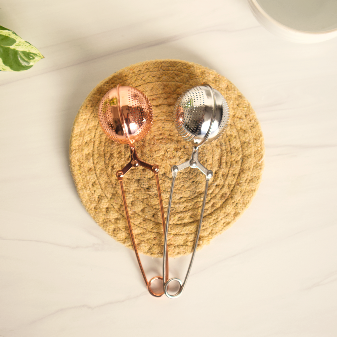 Two tea infusers, one copper and one silver, on a woven mat.