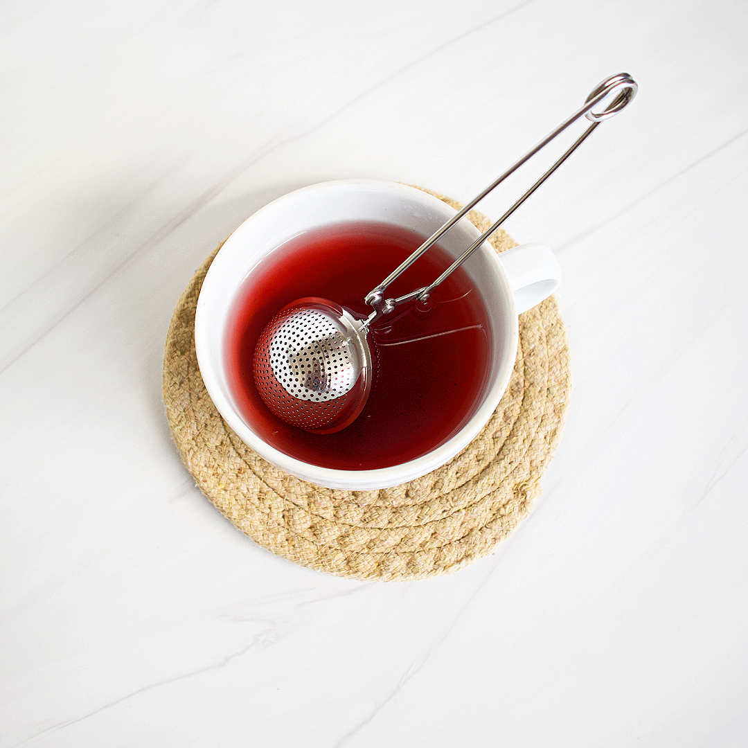 Tea cup with red liquid and a metal tea ball on a woven coaster.