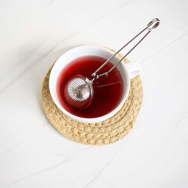Tea cup with red liquid and a metal tea ball on a woven coaster.