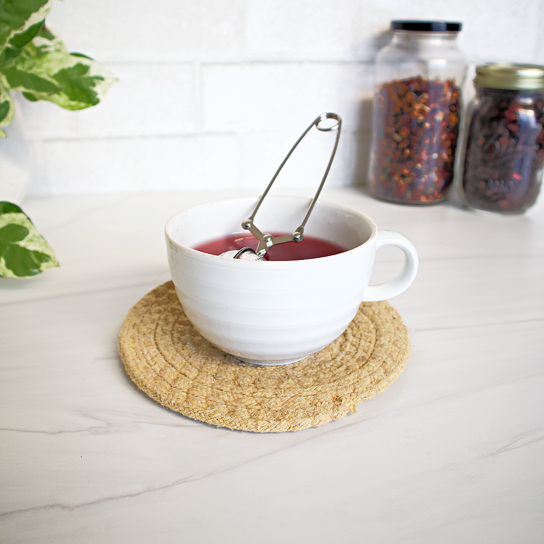 White mug with tea on a woven coaster on a light wooden surface with jars and a plant in the background.