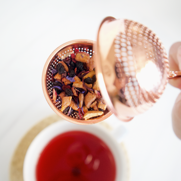 Copper tea infuser with dried herbs over a cup of tea on a white background