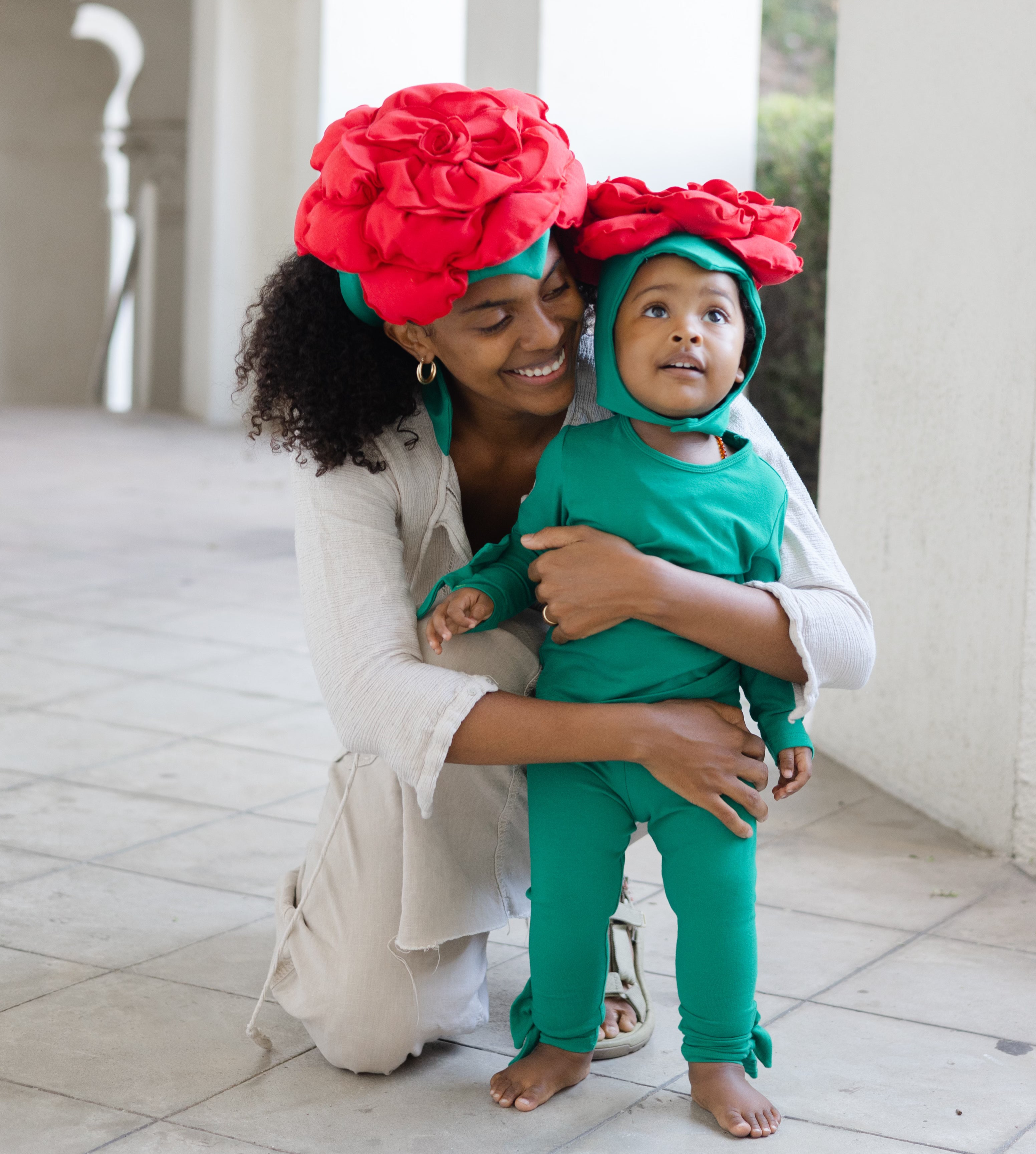 Woman holding a child both wearing green outfits with red flower headbands outdoors.