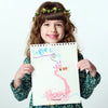 Child holding a drawing of a pink dragon with 'Sasha' written on it, wearing a floral crown.