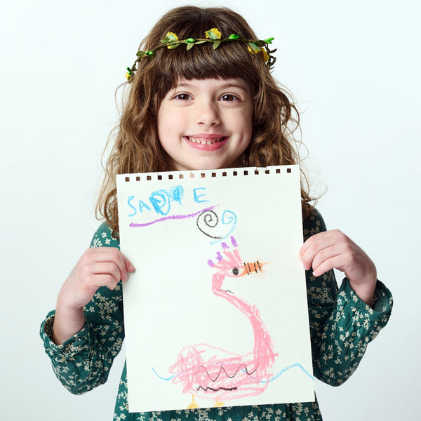 Child holding a drawing of a pink dragon with 'Sasha' written on it, wearing a floral crown.