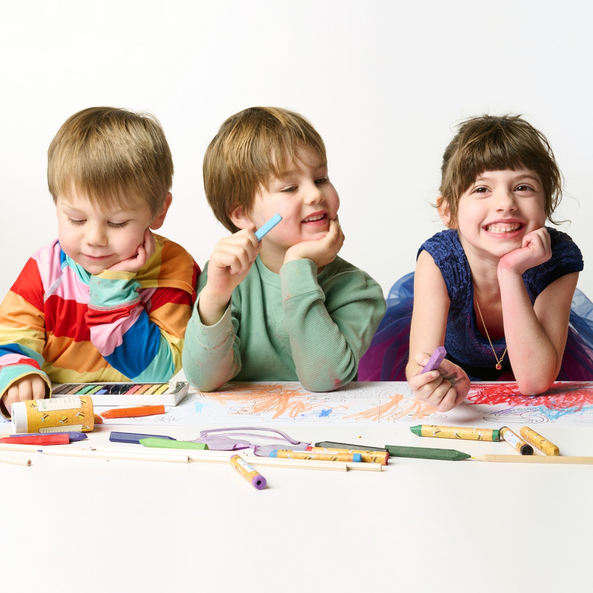 Three children sitting at a table with colorful crayons, smiling and engaged in an activity.