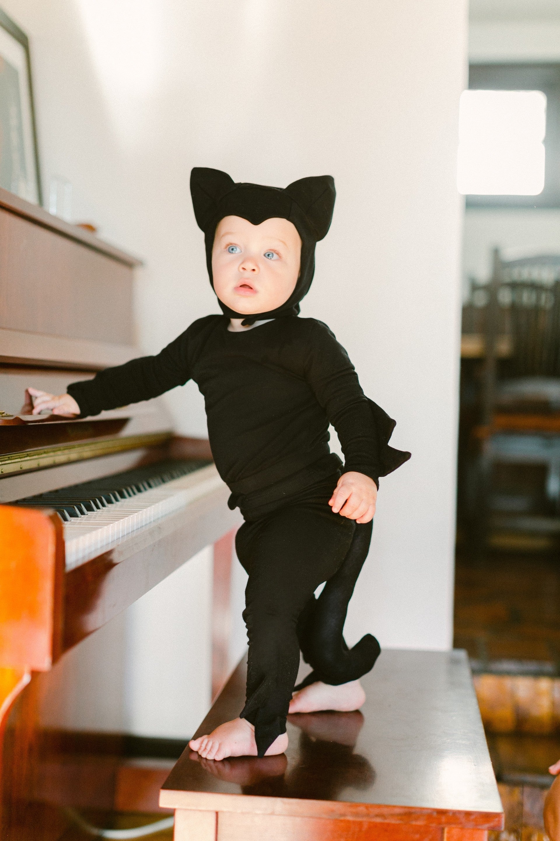 Child in a black cat costume standing on a piano stool next to a piano.