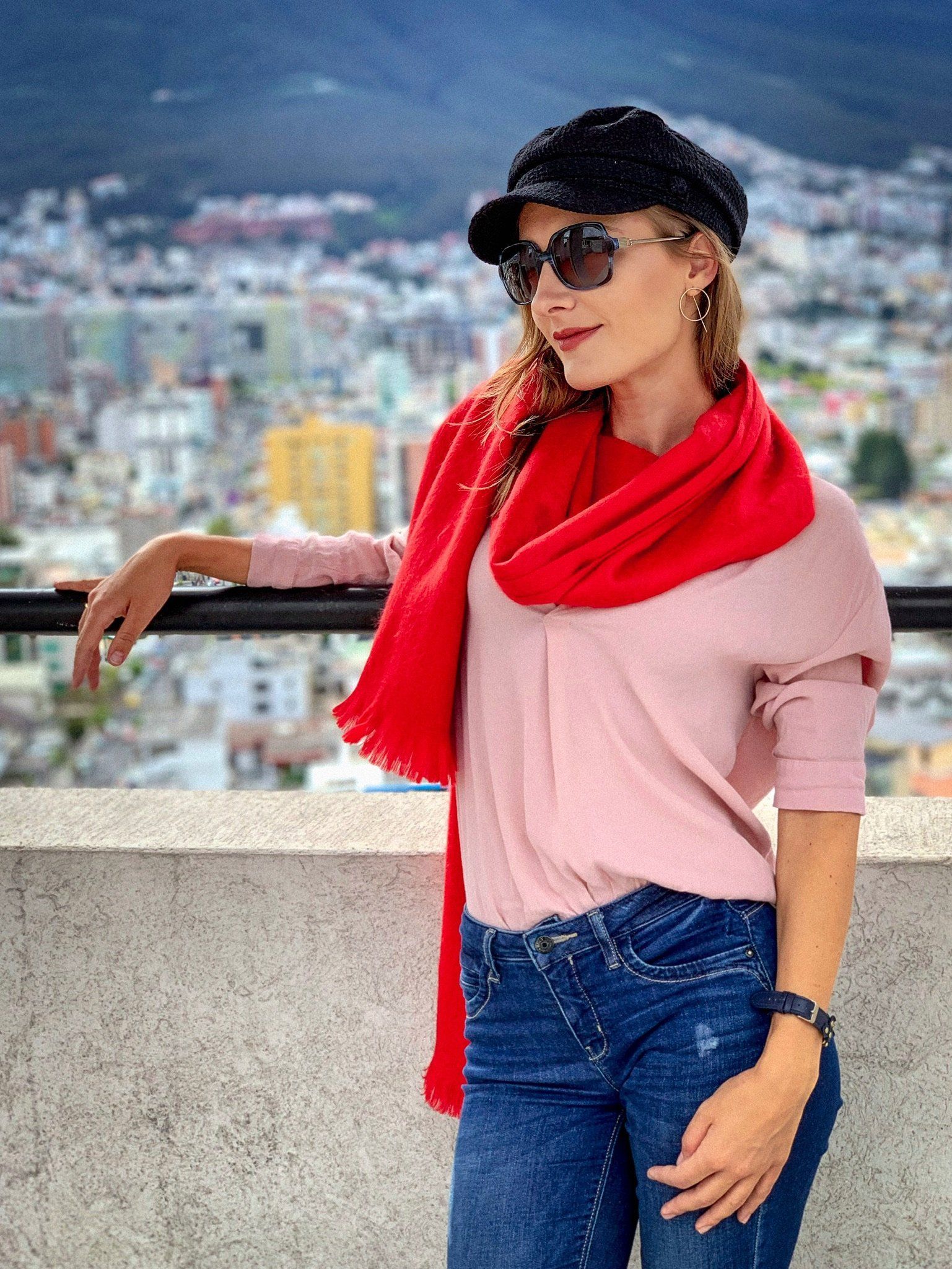Woman wearing a red scarf, pink top, and black cap with sunglasses, standing against a cityscape.