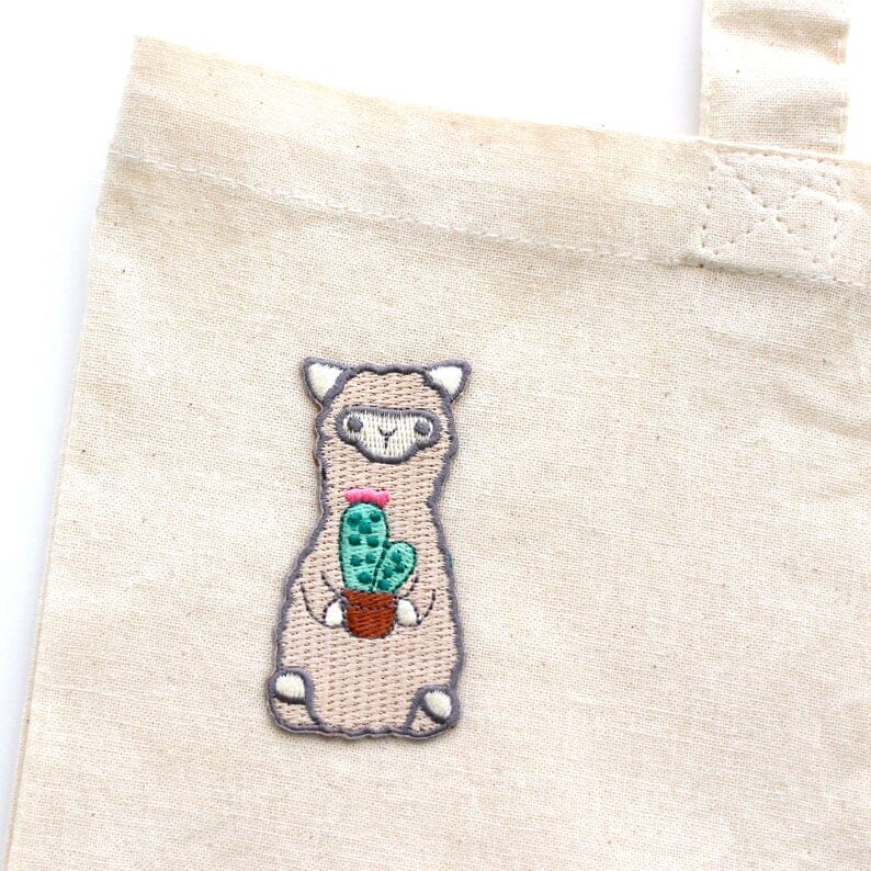 Embroidered llama holding a cactus on a beige fabric background