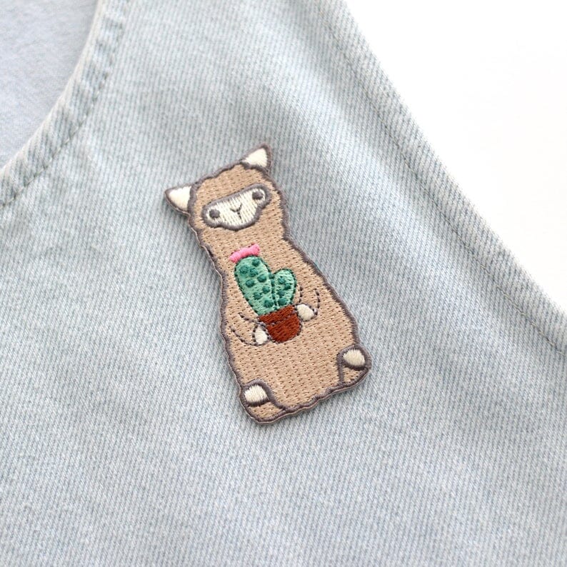 Embroidered patch of a llama holding a cactus on a denim fabric background