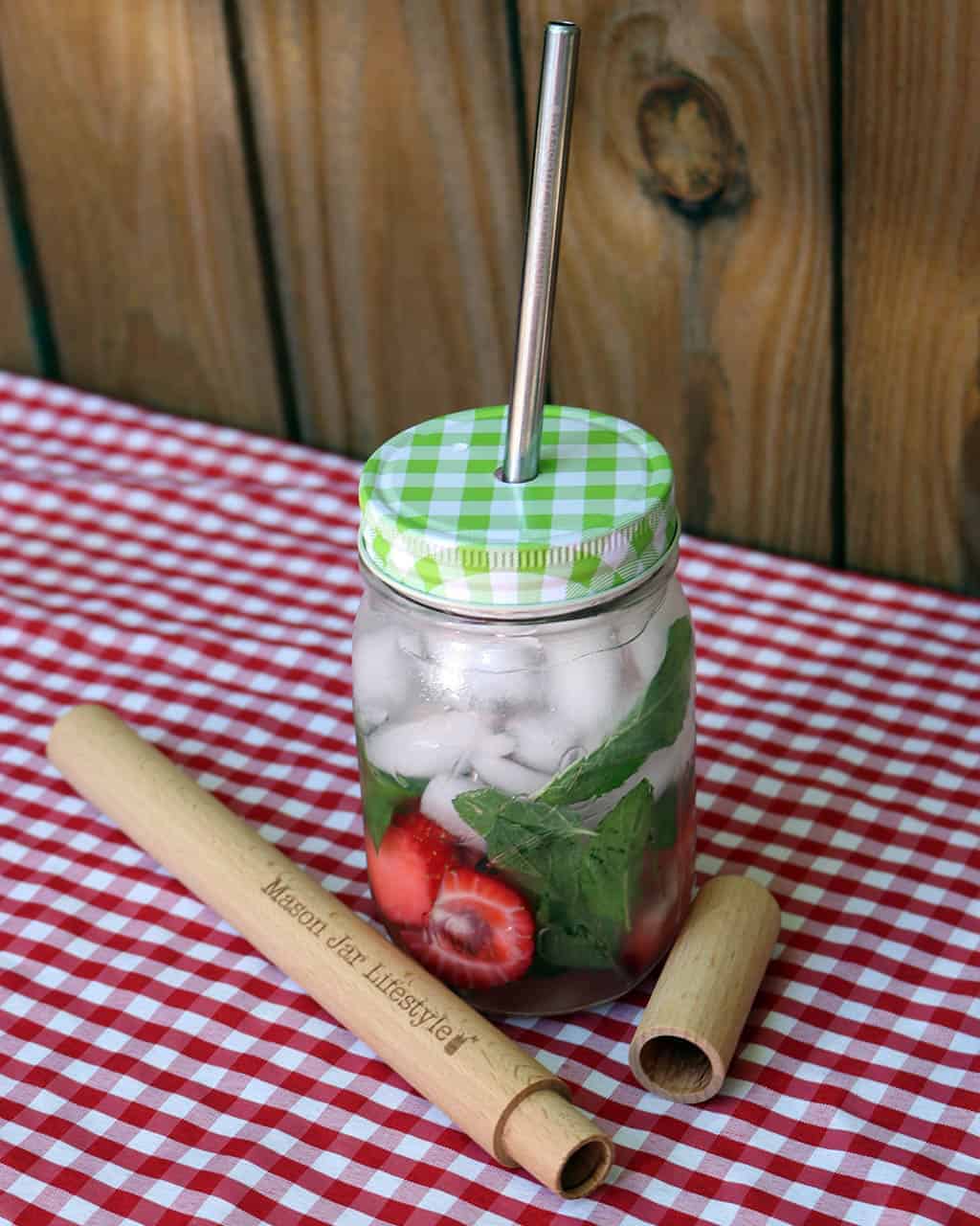 Mason jar with green lid, straw, and wooden stick on a red and white checkered tablecloth.