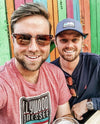 Two men posing together in front of a colorful wooden wall.