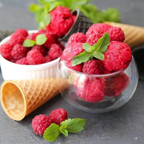 A clear double-walled thermo glass bowl filled with red raspberry sorbet, alongside a waffle cone and scattered raspberries on a dark surface.