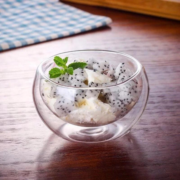 A clear double-walled vacuum sealed thermo glass bowl on a wooden surface, containing what appears to be a cold dessert or salad, garnished with a mint leaf.