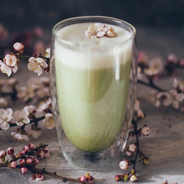 A clear double-walled Thermo Glass with a beverage inside, placed on a surface surrounded by cherry blossoms.