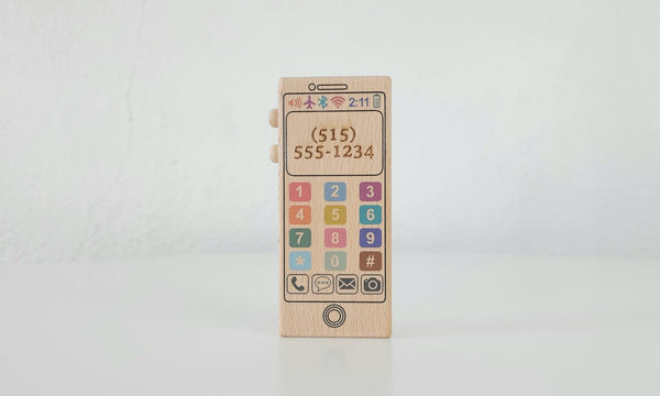 A colorful wooden toy phone made by Bannor Toys. The phone is made of maple wood and has a natural beeswax and flaxseed oil finish. It is a sleek design with numbers and letters, and it has a bell inside that makes a gentle noise when shaken. The phone is sitting on a white background.