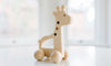 A photo of a wooden giraffe push toy. The giraffe is made from maple hardwood with cherry and walnut accents. It has wheels and a smooth, rounded design