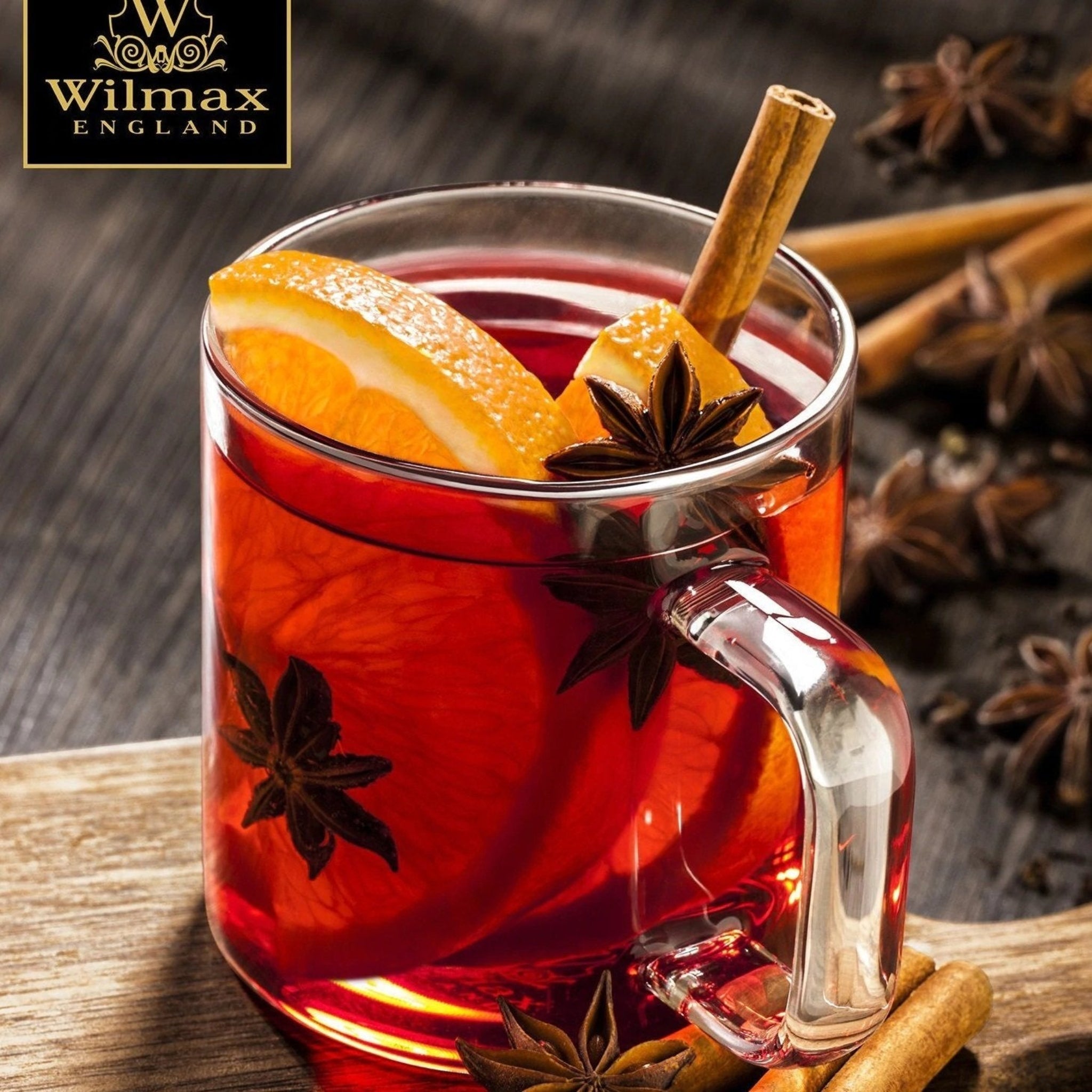 A clear glass mug with a handle, filled with a red beverage, alongside a cinnamon stick and an orange slice. The mug is placed on a dark surface, with whole spices around it.